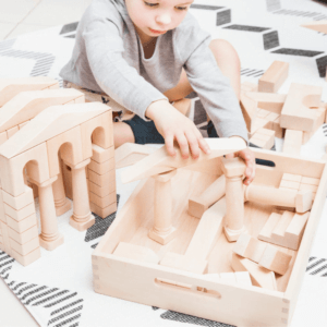 Child is playing classic building block set which includes regular basic block shapes and more complex ones such as arches, columns and triangular prisms.